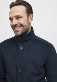 Mornarski vuneni kaput s podignutim ovratnikom, koji ima tri velika dugmeta i suptilnu teksturu. Dizajn je krojen i strukturiran.