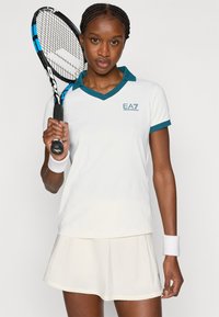 Άσπρο αθλητικό πόλο με τυρκουάζ λεπτομέρειες και V-λαιμόκοψη, συνδυασμένο με ανοιχτόχρωμη φούστα. Παίκτης κρατά ρακέτα τένις, με ορατό βραχιολάκι στον καρπό.