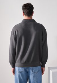 Maglione grigio scuro con tessuto strutturato, vestibilità comoda e polsini a costine. Indossato con jeans in denim azzurro chiaro, dal design semplice.