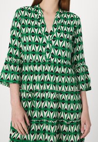 V-kauluksen, liehuvien hihojen ja kerroksellisten frillojen kanssa varustettu vihreä ja valkoinen kuvioitu mekko. Sisältää mustia ja vihreitä sävyjä geometrisilla muodoilla.