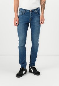 Man wearing slim-fit blue jeans and black sneakers with white stripes, standing against a plain white background.