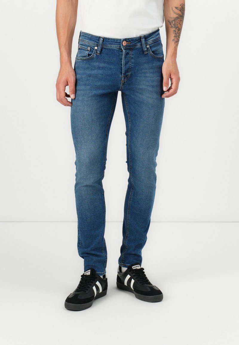 Man wearing slim-fit blue jeans and black sneakers with white stripes, standing against a plain white background.