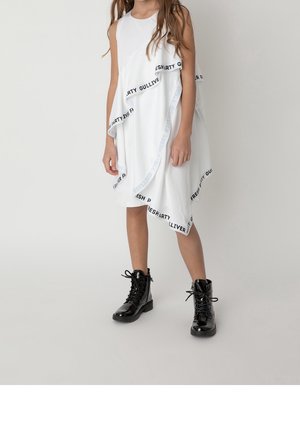 Hvid ærmeløs kjole med asymmetrisk kant og sorte brandingdetaljer, kombineret med glansfulde sorte snørestøvler. Glat stoftekstur.