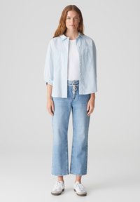 Lichtblauw geknoopt overhemd met opgerolde mouwen, witte onderhemd, hooggesneden licht gewassen jeans en witte sneakers met grijze zolen.