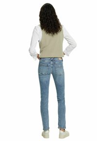 Ljust grön ribbad väst över en vit långärmad skjorta, i kombination med åtsittande ljusblå jeans och vita sneakers. Sedd från ryggen.