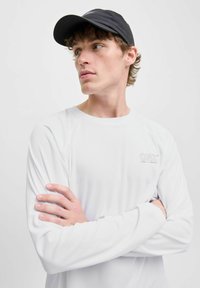 Wit sportshirt met lange mouwen van textuurstof, met een klein grijs logo op de linkerborst en een ronde halslijn. Een zwarte pet maakt de look af.
