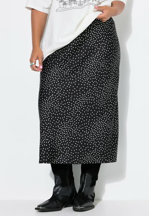 Personne portant une jupe midi noire à pois blancs, des bottes noires et un t-shirt blanc avec un motif graphique partiellement relevé par sa main.