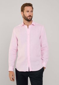 Camisa de manga larga rosa claro hecha de una tela texturizada, con un cierre de botones en el frente y un cuello clásico. Combinada con pantalones oscuros.