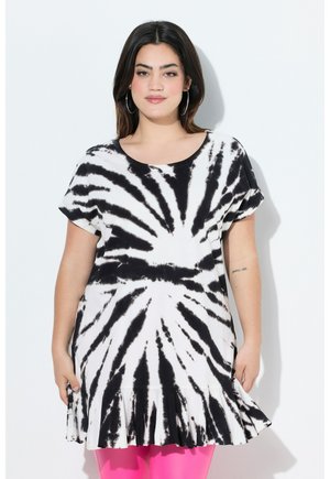 Frau trägt ein schwarz-weißes Batik-Kurzarmkleid mit gerafftem Saum und knallpinken Leggings, steht vor weißem Hintergrund.