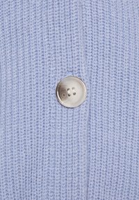 Gros plan d'un bouton deux trous gris nacré cousu sur un tissu tricoté texturé bleu pâle.
