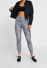 Veste zippée noire, brassière de sport noire et legging gris à imprimé léopard. Les baskets présentent un schéma de couleurs noir et blanc. Matière lisse et extensible.