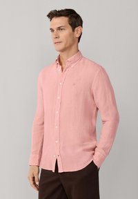 Homme portant une chemise rose à manches longues avec un petit logo brodé, associée à un pantalon brun foncé, debout contre un mur gris.