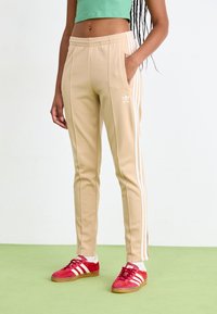 Beežid spordipüksid valgete triipudega, elastse vöökoha, küljetaskute ja adidas logo'ga. Komponeeritud punaste tossudega ja rohelise ülaosaga.