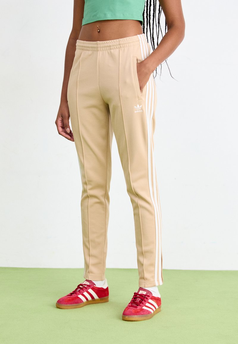Beežid spordipüksid valgete triipudega, elastse vöökoha, küljetaskute ja adidas logo'ga. Komponeeritud punaste tossudega ja rohelise ülaosaga.