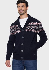 Merinotakki tummansinisellä värillä, jossa on huivikaulus ja Fair Isle -kuviointi punaisella ja valkoisella. Edessä kuusi suurta nappia ja ribbauskohdat hihoissa.