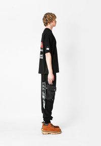 La camiseta negra de gran tamaño presenta un logotipo rojo y texto en la parte posterior. Los pantalones cargo negros tienen bolsillos laterales y detalles impresos en blanco. Botas marrones con cordones.