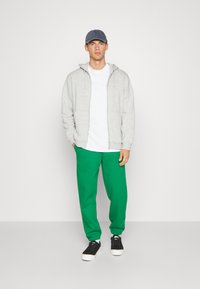 Grå zip-up hoodie, hvid t-shirt, grøn sweatpants med logo, sorte sneakers. En denim cap med skygge fuldender outfittet.