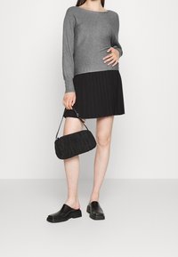 Donna che indossa un maglione grigio a righe, una gonna plissettata nera, scarpe slip-on nere, con in mano una piccola borsa nera su uno sfondo neutro.