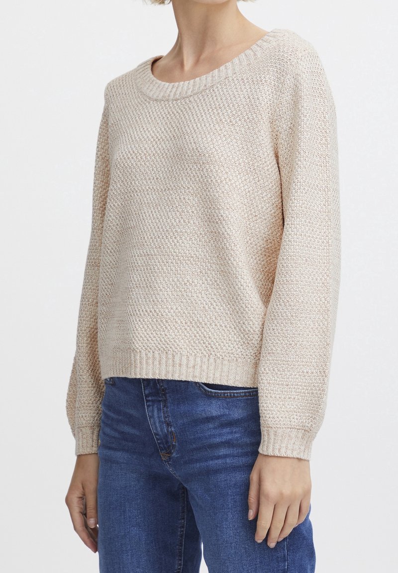Femme portant un pull en tricot beige texturé à manches longues et un jean bleu, sur un fond blanc uni.