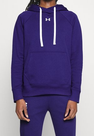 Osoba ubrana w fioletową bluzę z kapturem i dopasowane spodnie z białymi sznurkami oraz logo Under Armour na piersi i udzie.
