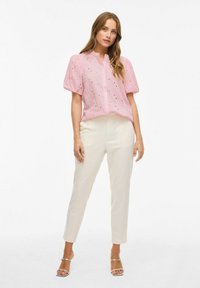 Chemise rose à manches bouffantes avec des découpes, associée à un pantalon tailleur blanc et des sandales à lanières argentées.
