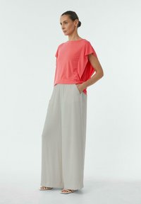Koralowy krótki top z krótkim rękawem o luźnym kroju, zestawiony z szerokimi jasnoszarymi spodniami z kieszeniami, noszony z białymi sandałami na paski.