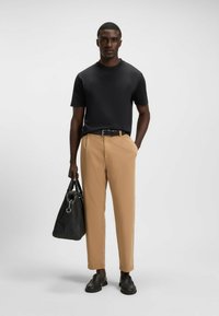 Schwarzes T-Shirt mit kurzen Ärmeln, beige Hose, schwarzer Gürtel und schwarze Schuhe. Trägt eine schwarze, strukturierten Handtasche. Einfaches und klares Design.