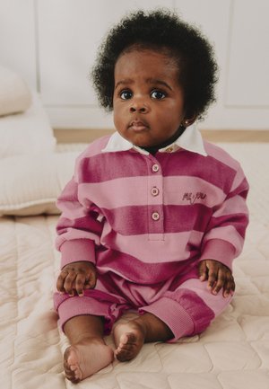 Baby med krøllet hår iført en pink og lilla stribet dragt siddende på et quiltet beige sengetæppe indendørs.