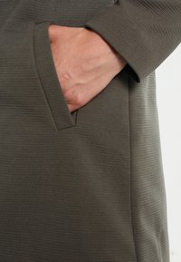 Tissu vert olive avec une texture côtelée, comportant une poche latérale. Une main repose à l'intérieur de la poche, mettant en avant un design à coupe décontractée.