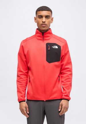 Młody mężczyzna w czerwonej kurtce na zamek błyskawiczny z czarną zamykaną kieszenią na piersi i logo The North Face, stojący na jednolitym białym tle.