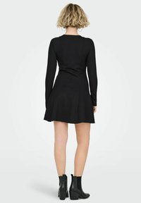 Kobieta o krótkich blond włosach ubrana w dopasowaną, czarną sukienkę z długim rękawem oraz czarne botki, stoi tyłem na jednolitym tle.