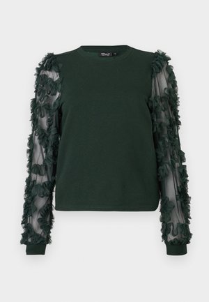 Haut à manches longues vert foncé avec un devant en tissu uni et des manches transparentes ornées d'appliqués floraux texturés.