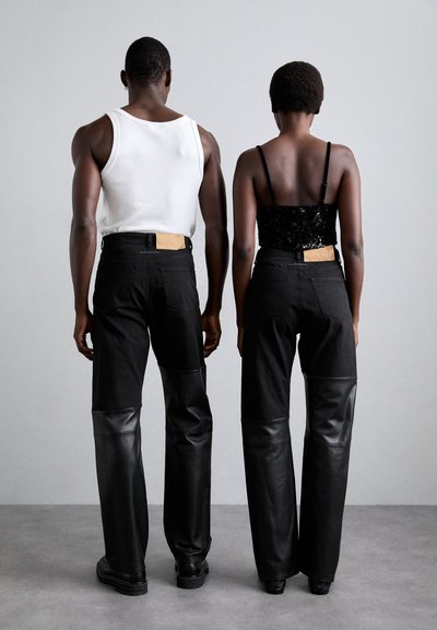 Black pants with leather panels at the bottom, worn by two models. One has a white tank top, the other a black strap top. Wood belt details.