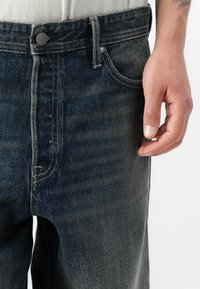 Donkergewassen denim jeans hebben een rechte snit, lichte stiksels en een metalen knoopsluiting. De stof heeft een gladde textuur met een lichte vervaging.