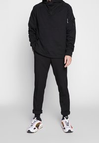 Černá mikina na zip s kapucí a dlouhými rukávy, volný střih, v kombinaci s černými zúženými kalhotami a bílými teniskami s oranžovými a černými detaily.