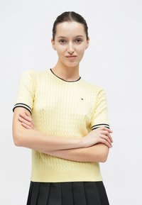 Κίτρινο πλεκτό τοπ με κοντά μανίκια που διαθέτει υφασμάτινο μοτίβο, μαύρα ριμπ μανσέτες και μικρή κεντημένη λογότυπο στο στήθος.