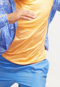 Tenue de sport comprenant une veste bleu à motif, un haut rayé orange et un short bleu à design géométrique. Les matériaux semblent légers.
