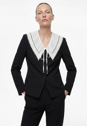 Vrouw gekleed in een zwart pak met een grote, witte kraag met kantafwerking, vastgebonden aan de voorkant, staande met handen in de zakken tegen een effen achtergrond.