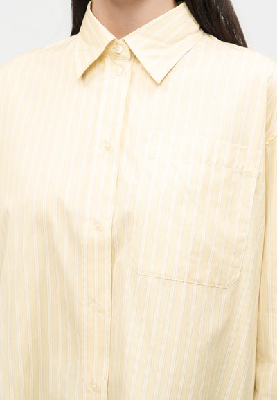 Γυναίκα που φοράει ανοιχτό κίτρινο πουκάμισο με λευκές κάθετες ρίγες και τσέπη στο στήθος, εμφανιζόμενη από τον λαιμό έως το μέσο του κορμού.