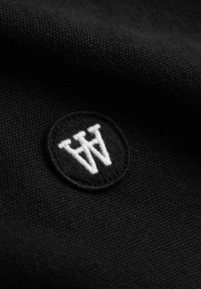 Tissu noir à surface texturée avec un patch circulaire brodé. Le patch présente un logo blanc stylisé composé de deux lettres "W".