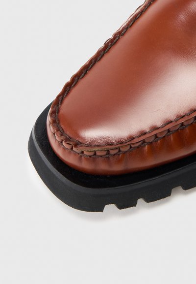 Brązowy skórzany but o gładkim wykończeniu, z kontrastowymi przeszyciami i czarną gumową podeszwą z bieżnikiem zapewniającym przyczepność. Zaokrąglony kształt palca.