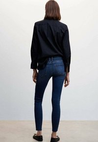 Woman standing with back turned wearing dark blue skinny jeans, black button-up shirt, and black ballet flats on a plain background.