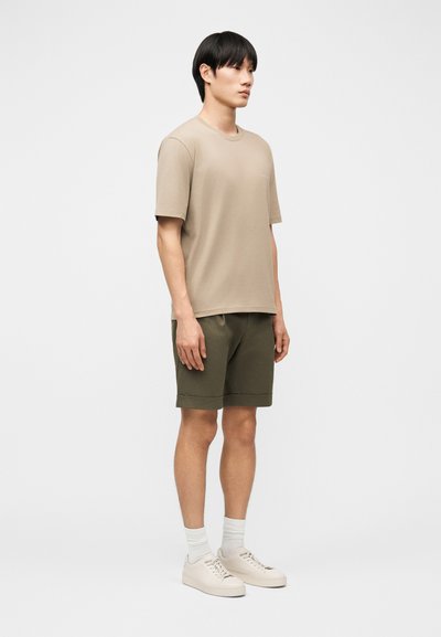 Άνδρας που στέκεται φορώντας μπεζ πουκάμισο με κοντά μανίκια, χακί πράσινα σορτς, λευκές κάλτσες και μπεζ αθλητικά παπούτσια σε ένα απλό ανοιχτό γκρι φόντο.