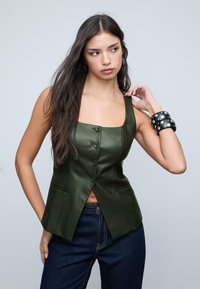 Top in satin verde oliva con scollatura quadrata, bottoni frontali e due tasche; abbinato a jeans in denim blu scuro e bracciali neri.