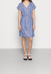 Helles blaues Kleid mit kurzen Ärmeln, weißen vertikalen Streifen, V-Ausschnitt, tailliertem Bund und lockerem Schnitt. Kombiniert mit weißen Sandalen.