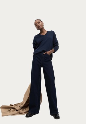 Navy V-neck sweater paired with wide-leg dark denim trousers. A beige fabric drapes on the floor, and black shoes complete the outfit.