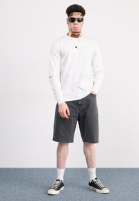 Camisa blanca de manga larga, pantalones cortos de mezclilla grises, zapatillas negras con suelas blancas y gafas de sol. El modelo está de pie sobre una alfombra azul con un fondo simple.