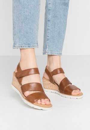 Fødder iført brune læder kilehælssandaler med ankelstropper kombineret med lyseblå cropped jeans på en hvid gulv.
