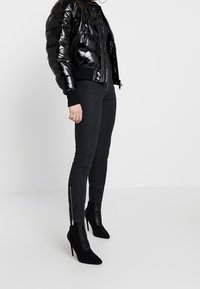 Svart blanka bomberjacka, åtsittande svarta jeans med siddragkedjor och svarta högklackade ankelboots, allt i en stilren, monokromatisk stil.