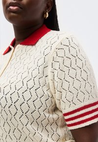 Temnopolta oseba, oblečena v kremasto pleteno polo majico z rdečim ovratnikom in kratkimi rokavi z rdečimi črtami, ter zlatimi uhančki v obliki obročev.